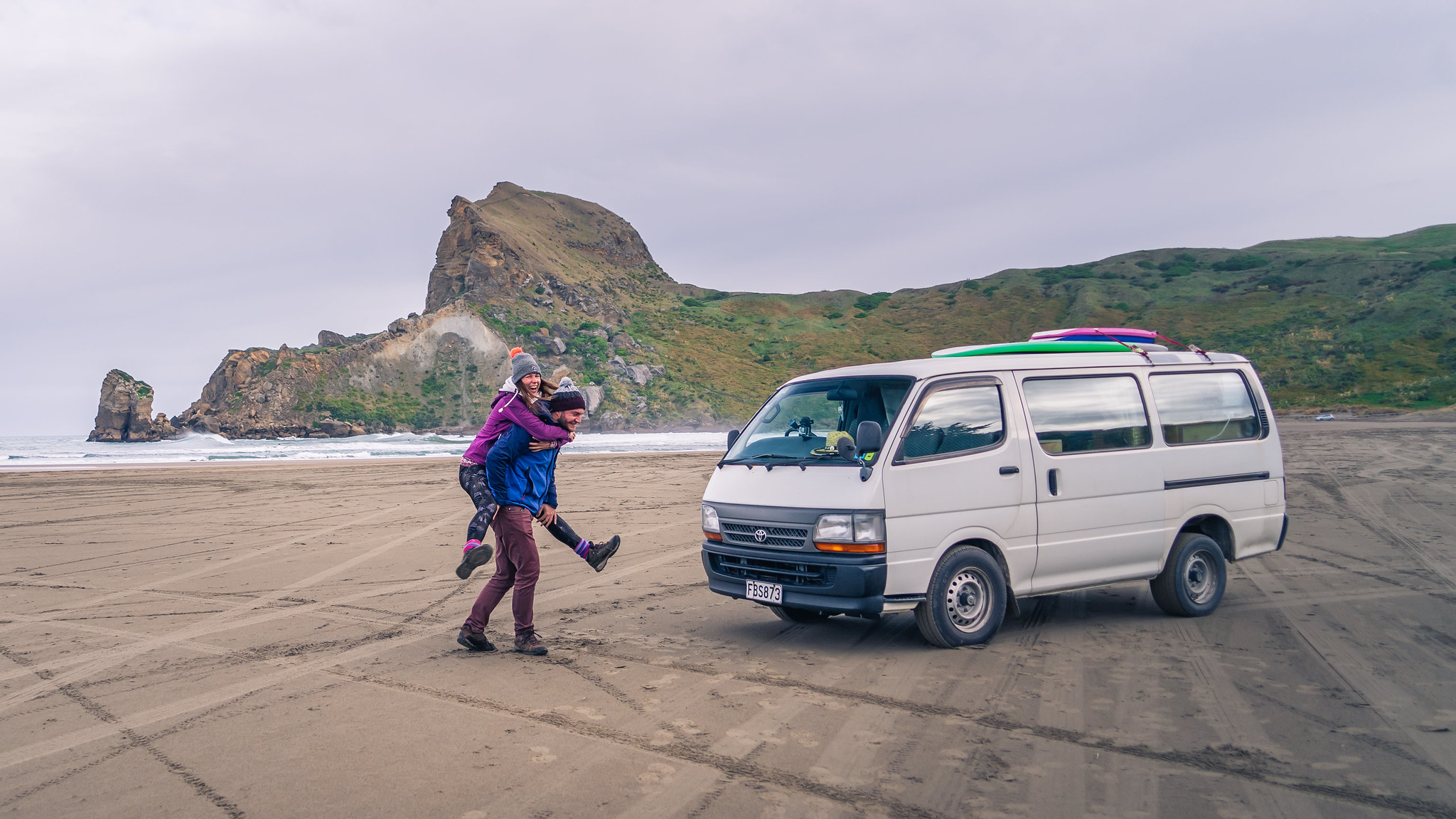Stan and Mishka laughing beside Vanilla, their original campervan, on a New Zealand beach